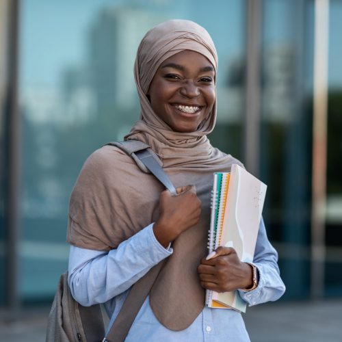 College Scholarships for Muslim Students. Happy Black Islamic Woman With Backpack And Workbooks Posing Outdoors At Campus, Resting After Classes, Wearing Hijab, Looking And Smiling At Camera