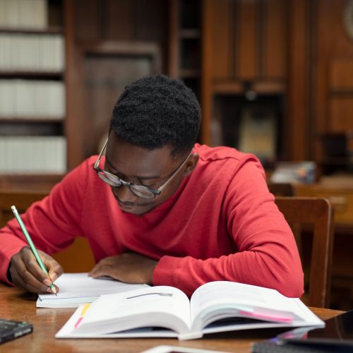University student wearing spectacles studying in library. Young african man taking notes from book while sitting in the library. Focused casual guy writing notes during homework.